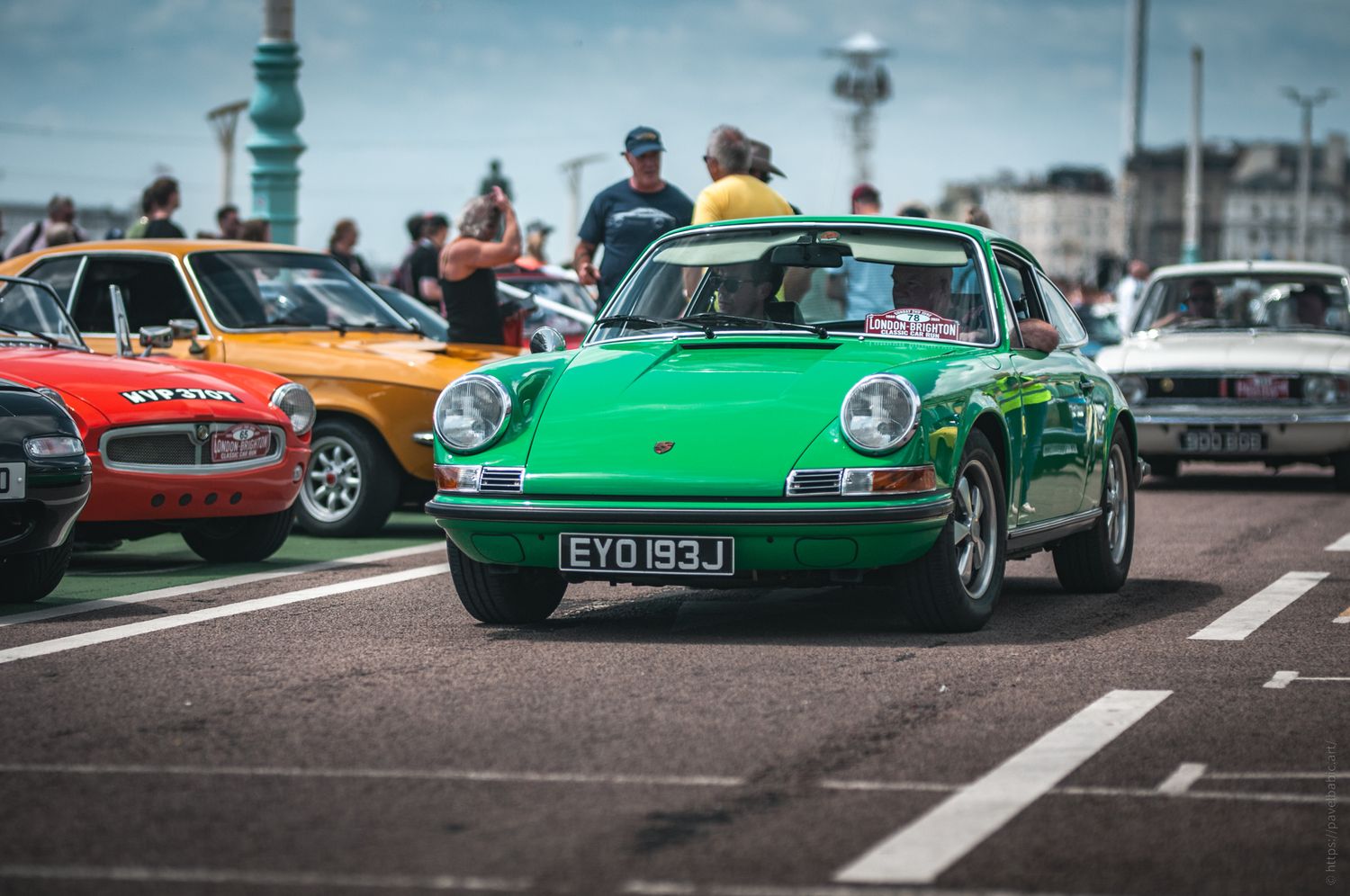 A vibrant green classic Porsche drives past a crowd at a car show, surrounded by colorful vintage vehicles on a sunny day.