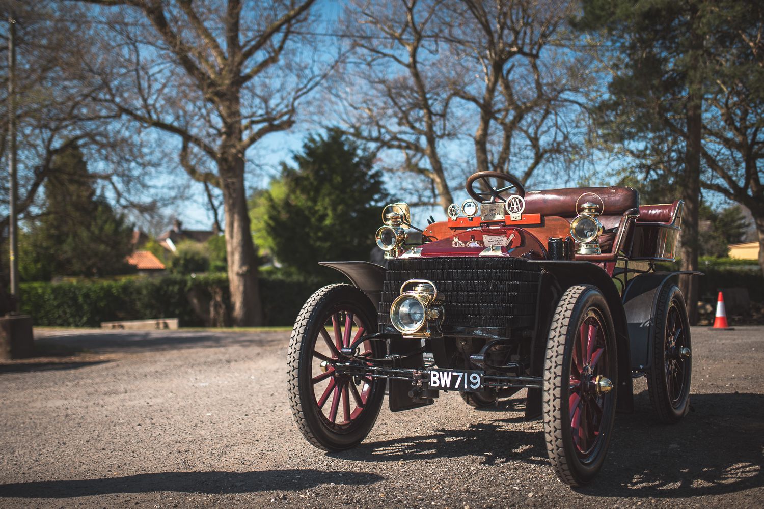 A vintage car with a red and black design, classic headlights, and wooden wheels parked under trees on a sunny day.
