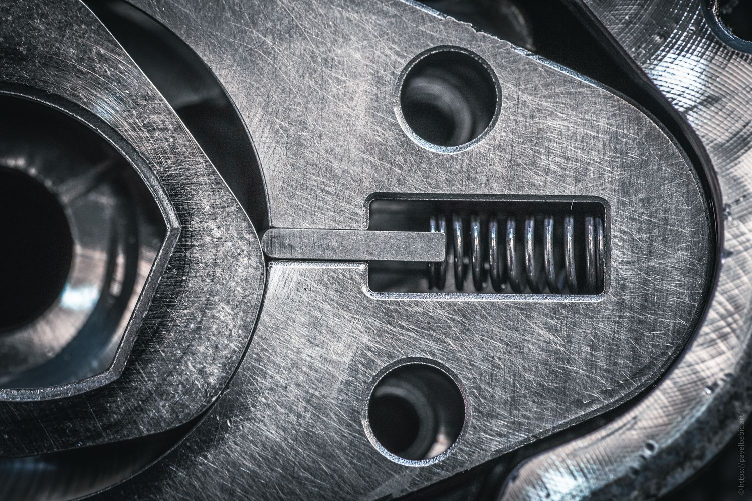 Close-up of a metal mechanical part showing a coiled spring, screws, and precision-engineered surfaces.