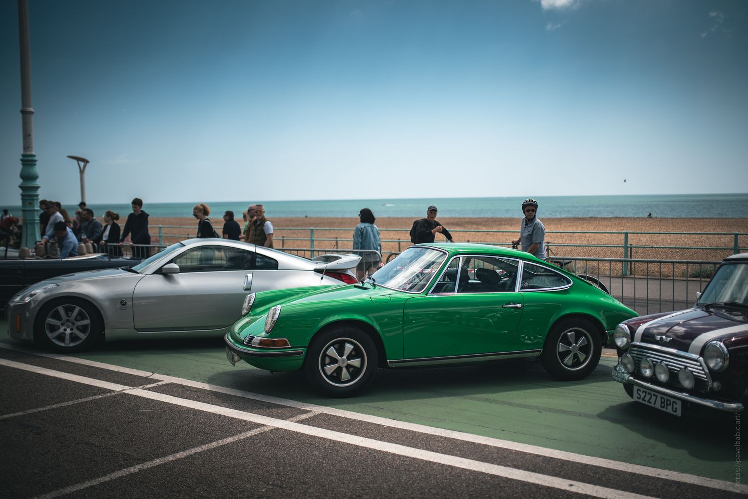 A vibrant green classic car parked by the beach, flanked by a silver coupe and a black mini, with people strolling nearby.