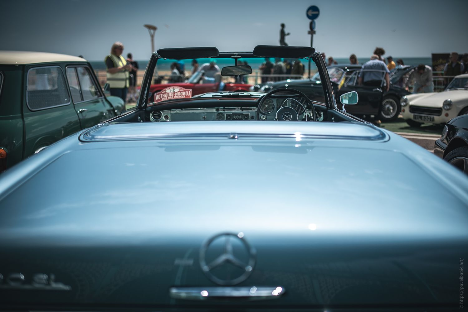 View of a vintage blue convertible Mercedes, with a busy seaside background and classic cars on display. Sunlit, nostalgic atmosphere.