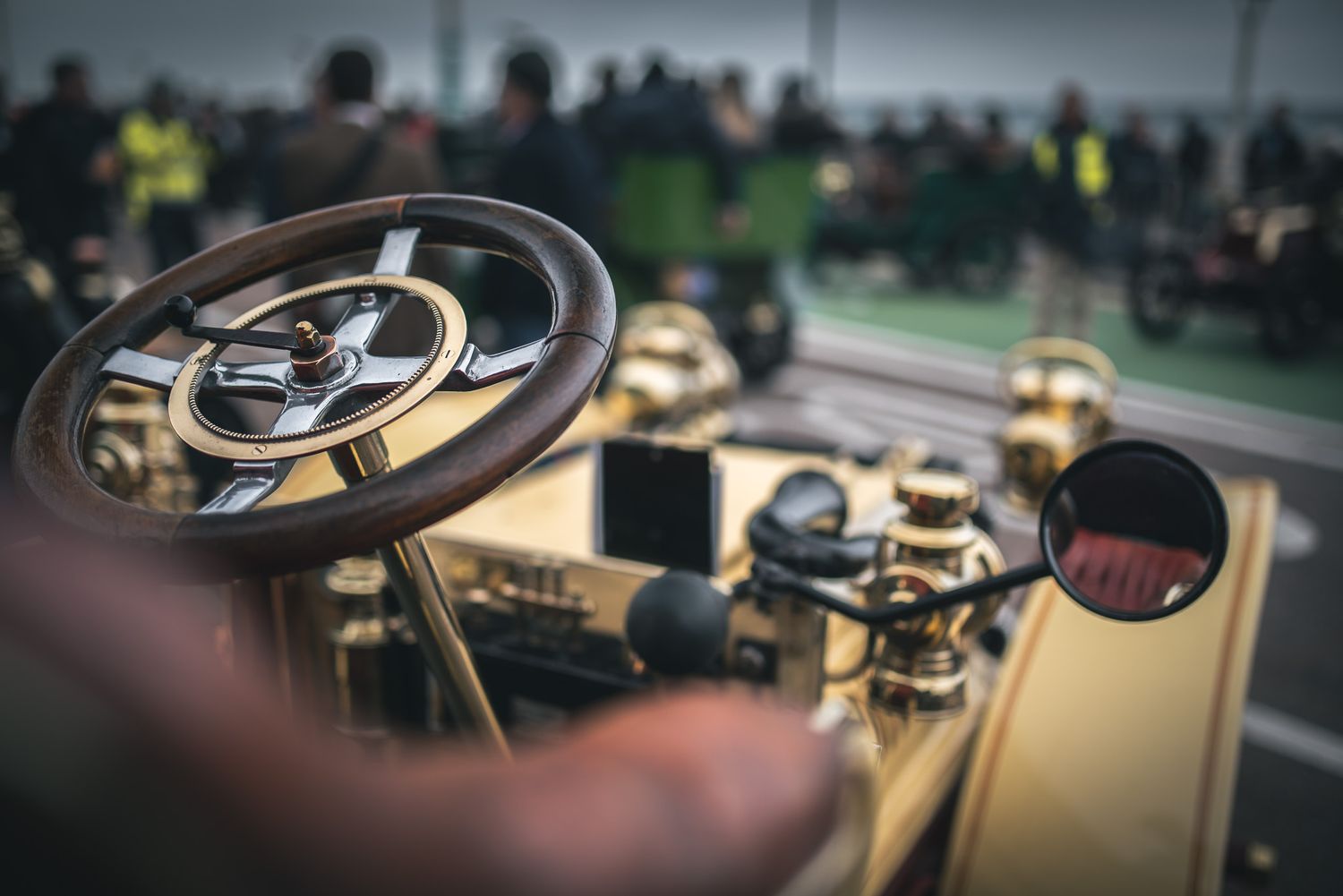 Close-up of a vintage car's wooden steering wheel and dashboard, with a blurred crowd in the background, creating a nostalgic and classic at