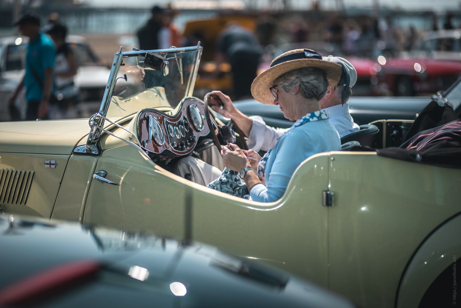 A couple sits in a vintage yellow convertible, with the woman wearing a straw hat, while looking at a phone amidst classic cars.