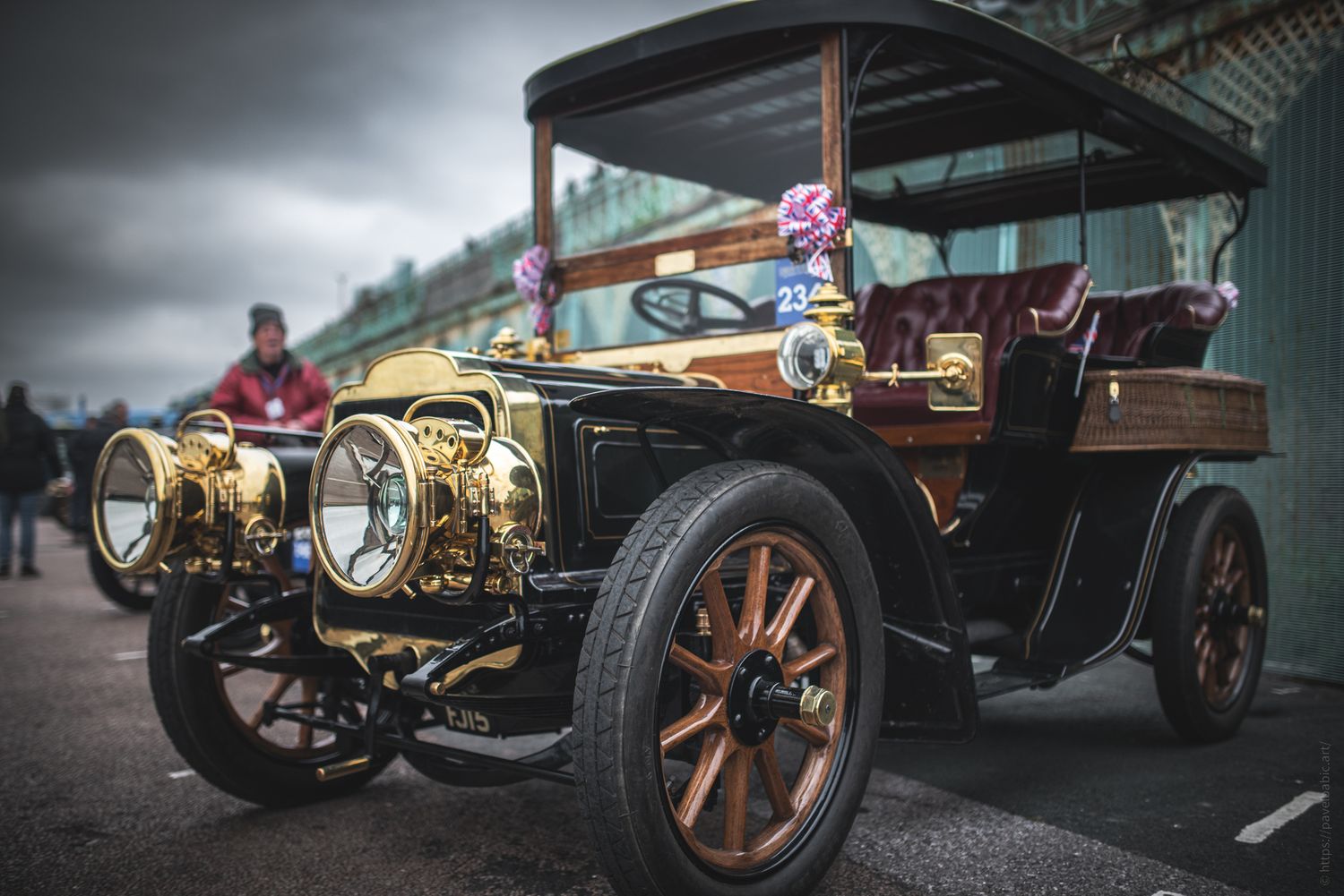 A vintage brass and black automobile with wooden accents, parked outdoors with people in the background, showcasing classic design.