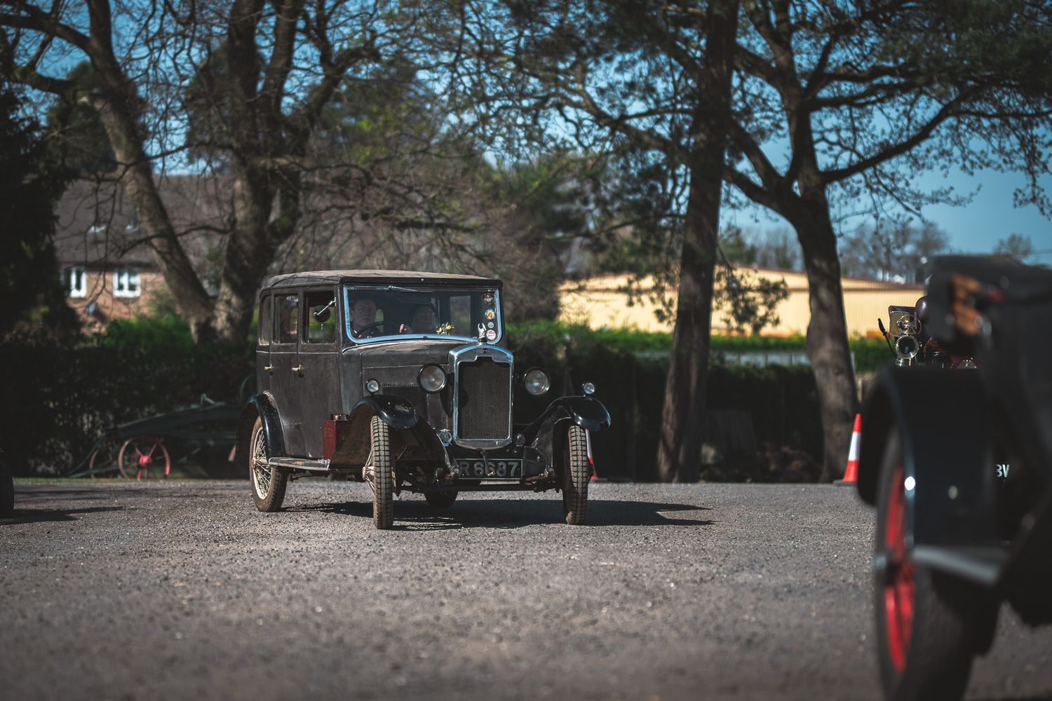 A vintage car, possibly from the 1930s, drives through a gravel area surrounded by trees and distant buildings on a sunny day.