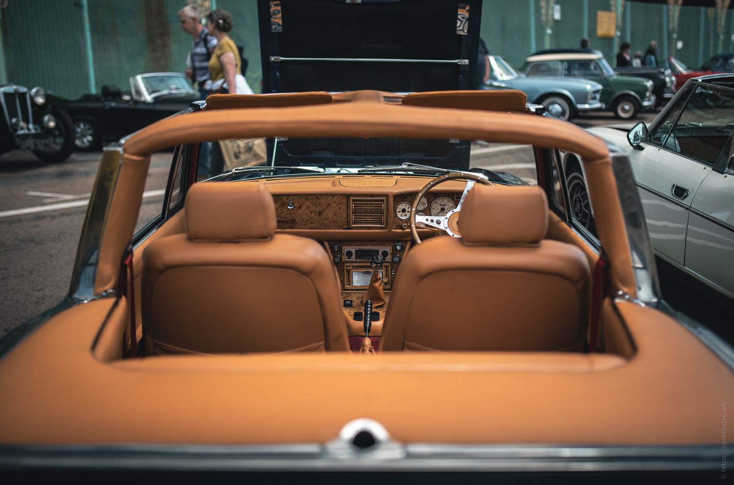 View from the backseat of a classic convertible car, featuring tan leather seats and a vintage dashboard, with other cars in the background.