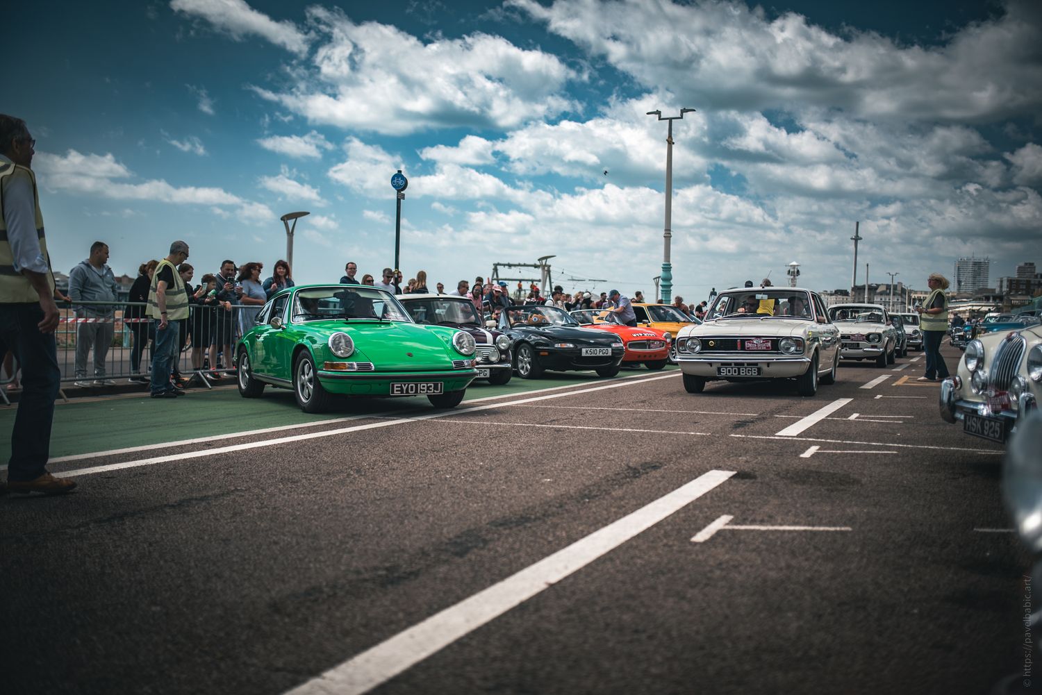 A vibrant car show with classic cars, including a green sports car, along a busy waterfront street, under a blue sky. Crowds gather to admir