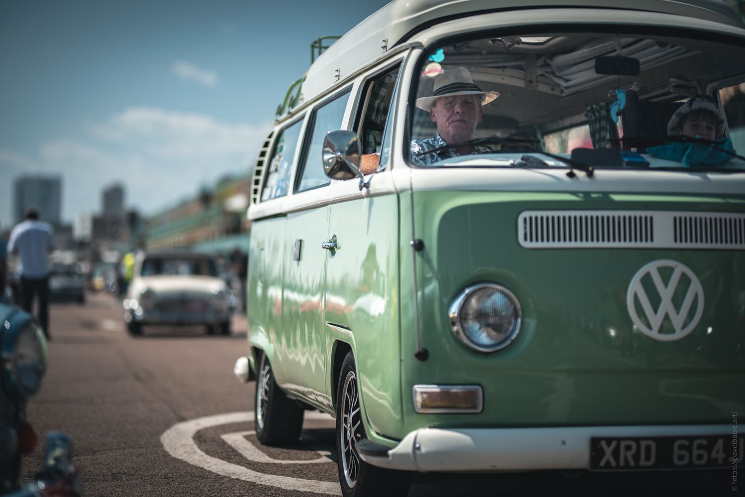 A classic green Volkswagen van parked in a busy area, with a driver visible inside, surrounded by other vintage cars.