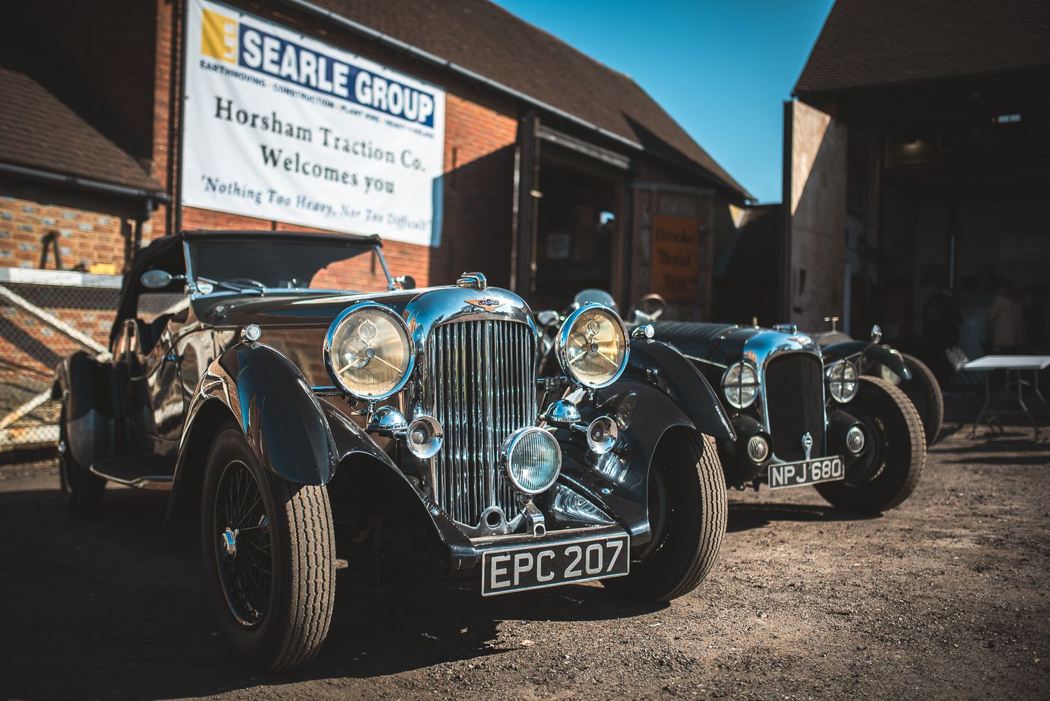 Two vintage cars parked in front of a barn, with a Searle Group banner welcoming visitors in the background. Bright sunny day.