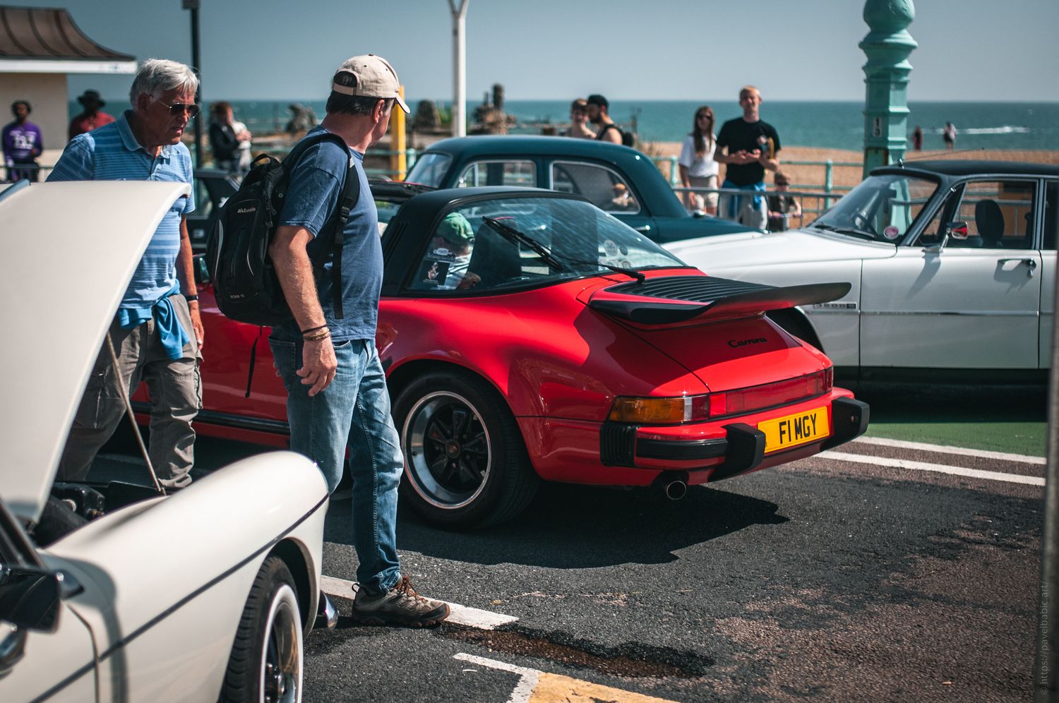 A red sports car parked by the beach, with people admiring it and classic cars in the background on a sunny day.