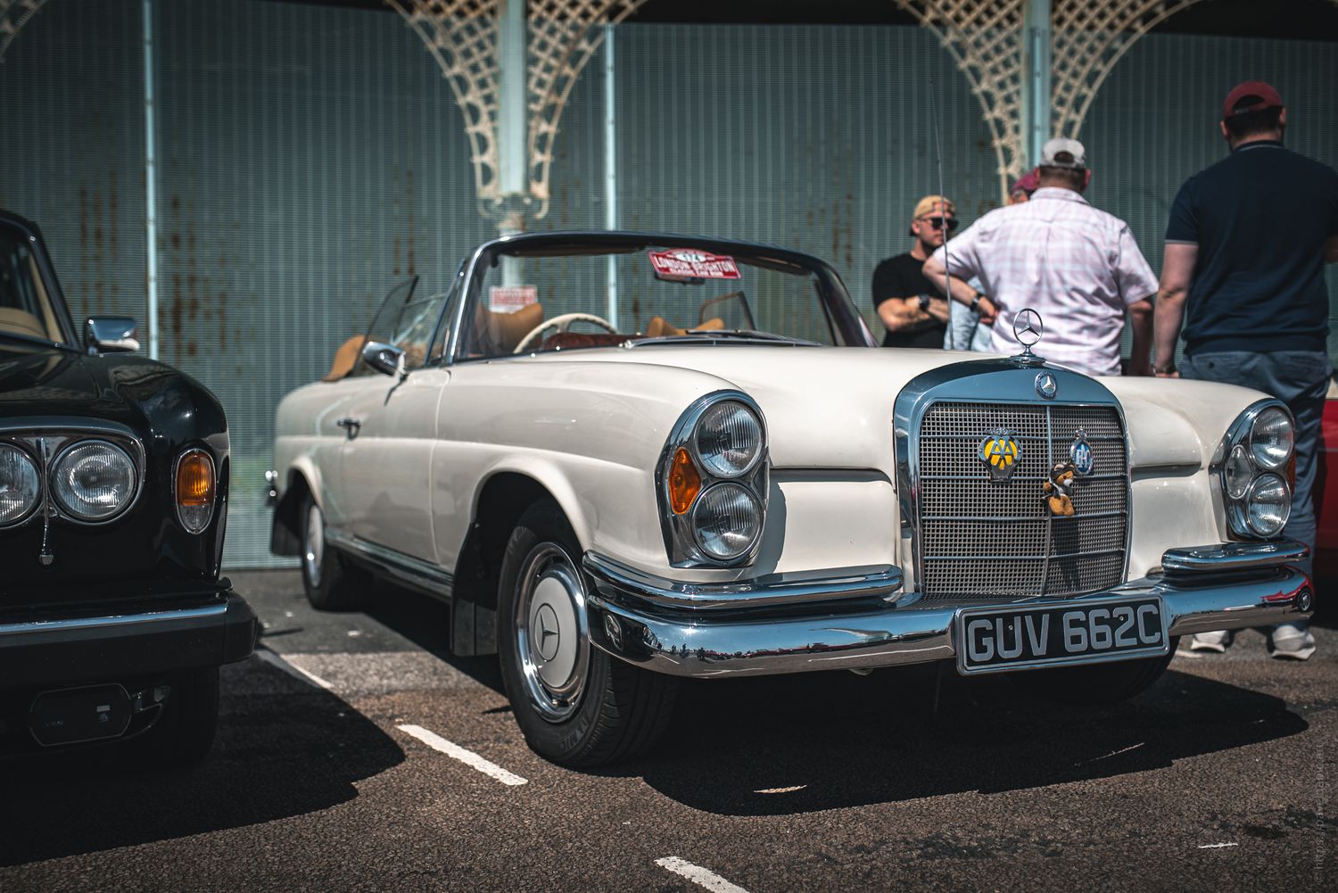 A vintage white convertible Mercedes-Benz parked beside a dark car, with people chatting in the background on a sunny day.
