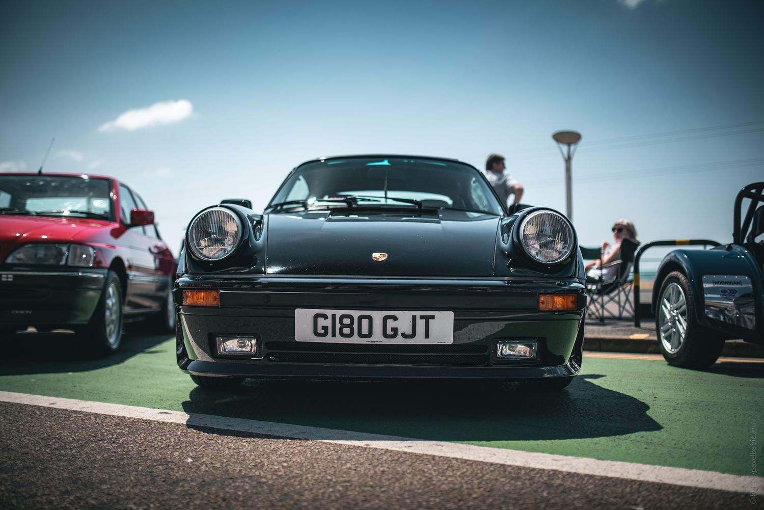 A sleek black sports car parked on a sunny day, flanked by a red vehicle and another sports car, with people visible in the background.