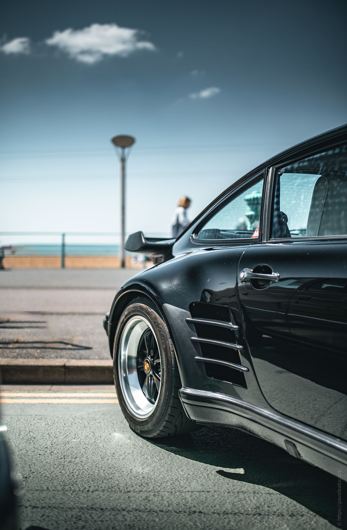 A sleek black sports car parked by the seaside, showcasing its stylish profile and alloy wheels against a clear blue sky.