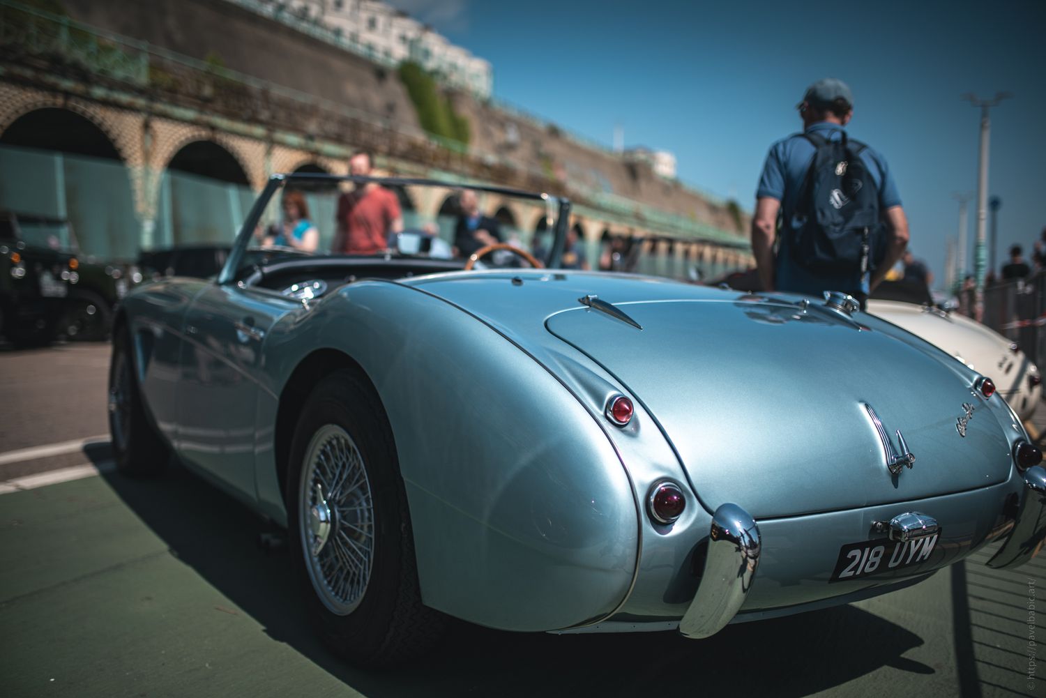A sleek, vintage silver convertible car parked on the street, with a blurred crowd and urban backdrop in bright daylight.