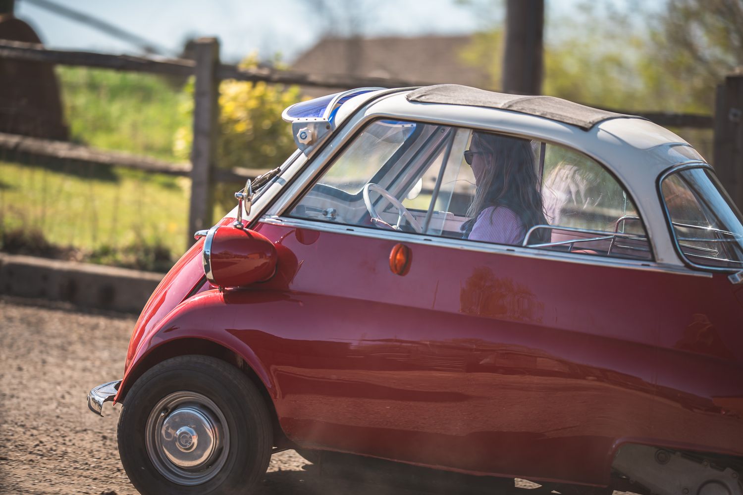 A woman driving a vintage red Isetta microcar, with a scenic background of greenery and a wooden fence on a sunny day.