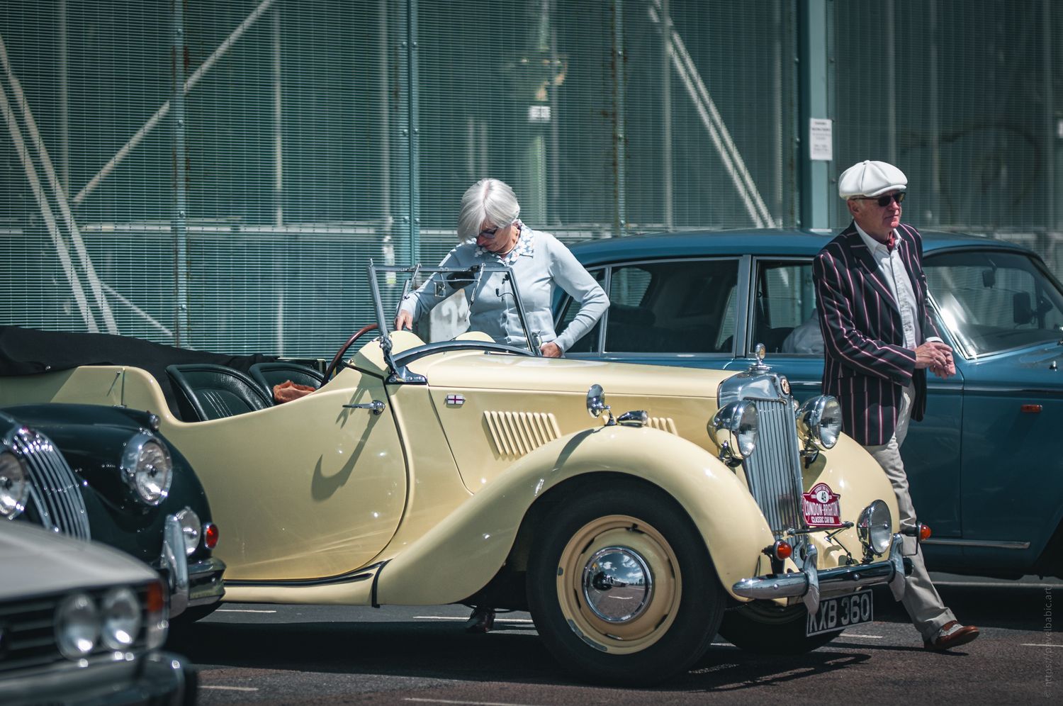 A classic yellow convertible car with two elderly people, a man in a striped jacket and a woman adjusting her shirt, standing nearby.