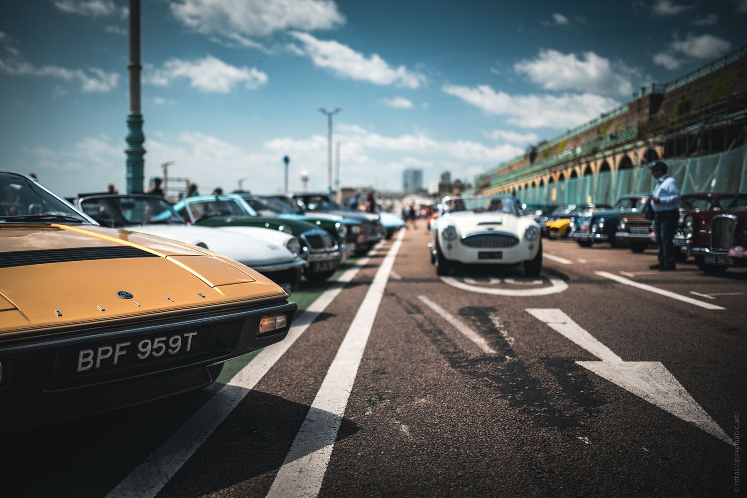 A sunny scene of classic cars lined up along a seaside promenade, with a clear blue sky and vintage architecture in the background.