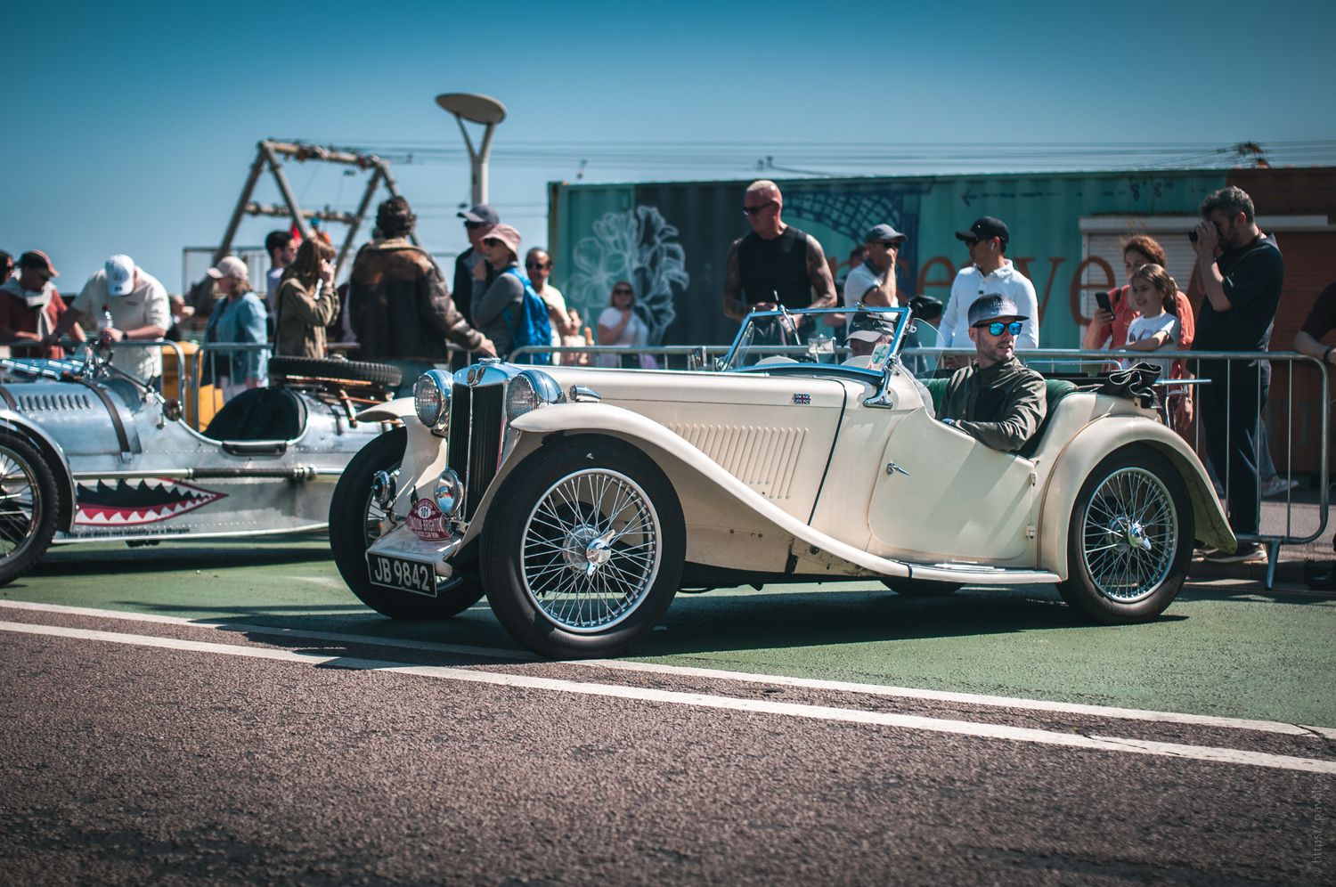 A vintage beige convertible car is parked at a car show, surrounded by onlookers admiring classic automobiles under a clear blue sky.