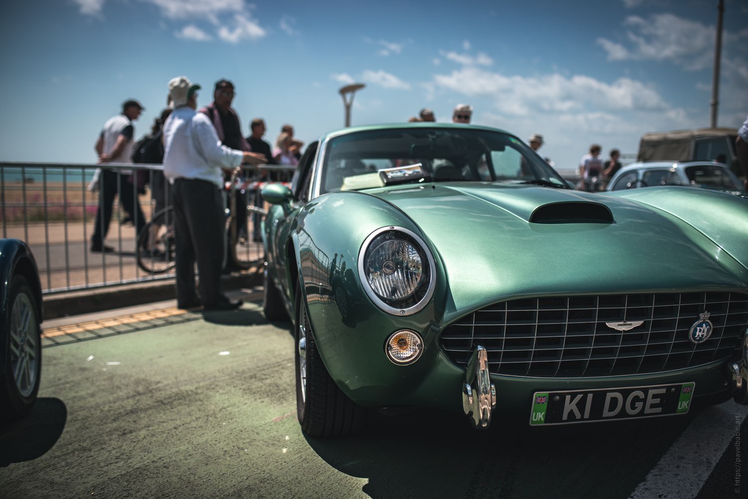 A sleek green vintage car parked near a beach, with people chatting in the background under a clear blue sky.