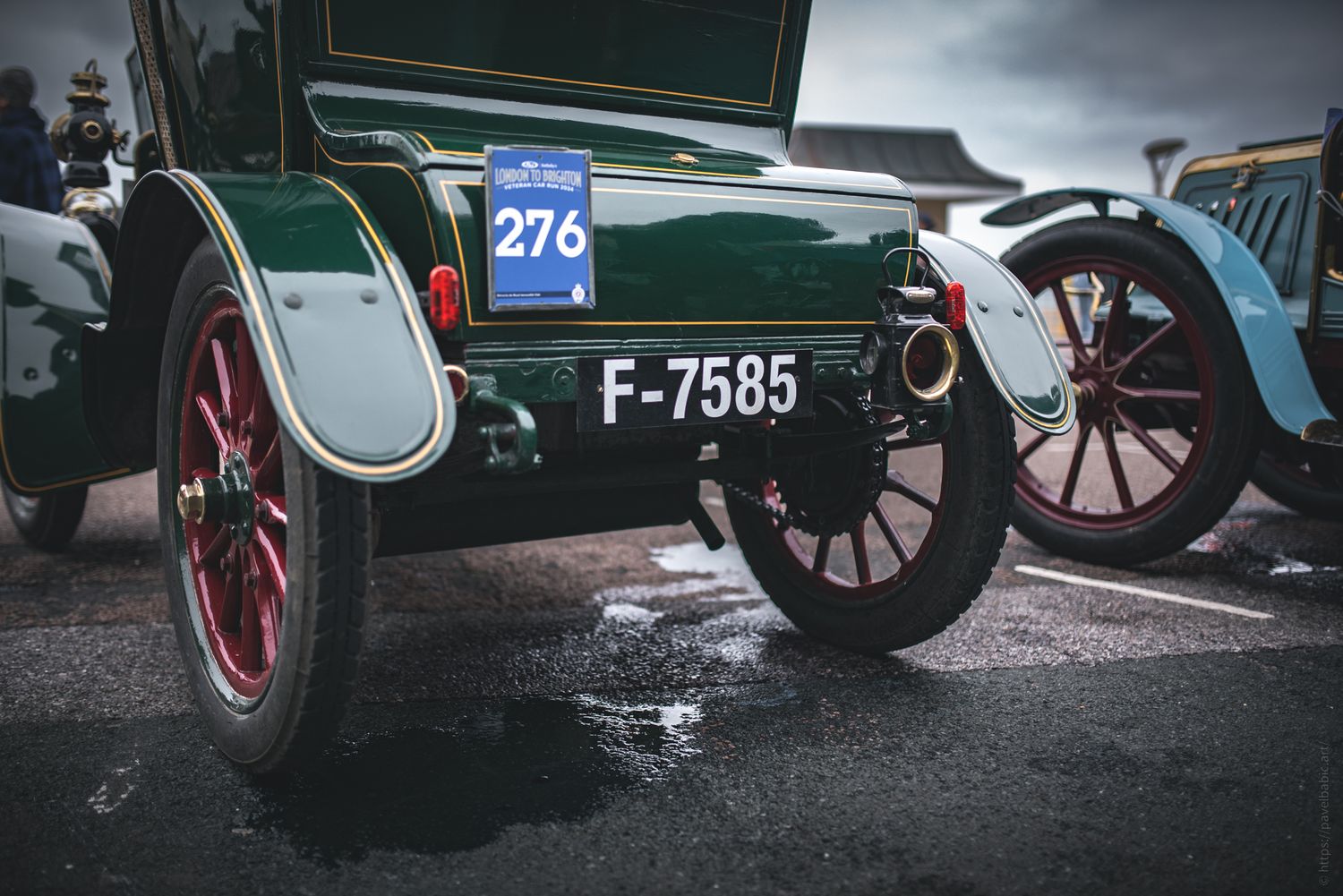 Close-up of vintage green car's rear wheel and license plate, showcasing classic design and details. Dark, overcast sky in background.
