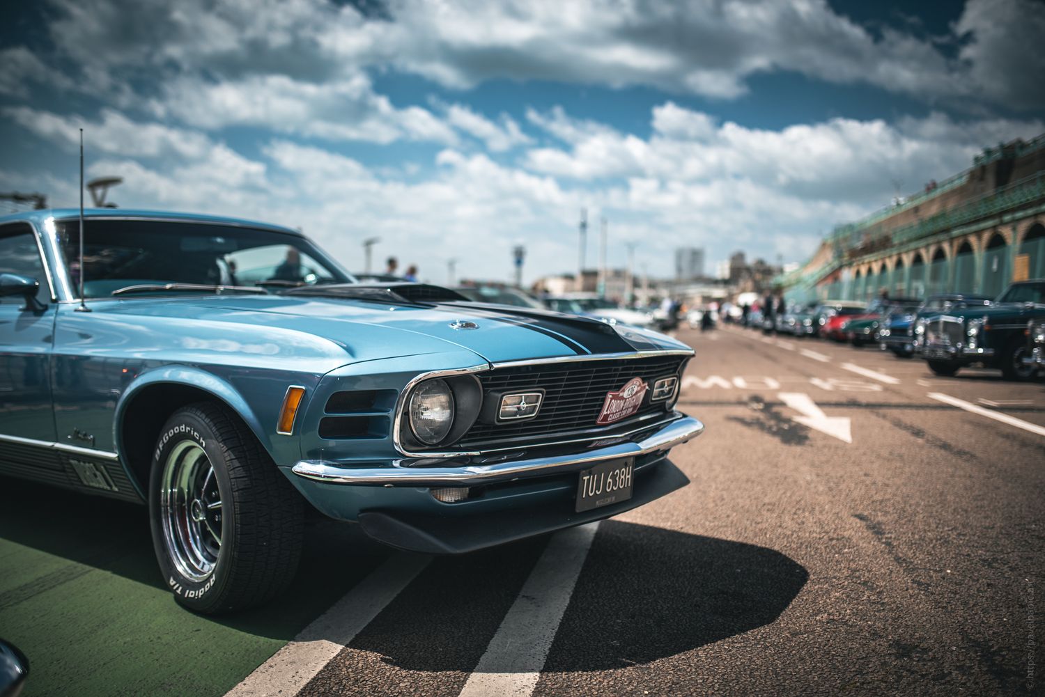 A classic blue 1970 Mustang parked in a scenic outdoor setting with other vintage cars in the background under a cloudy sky.