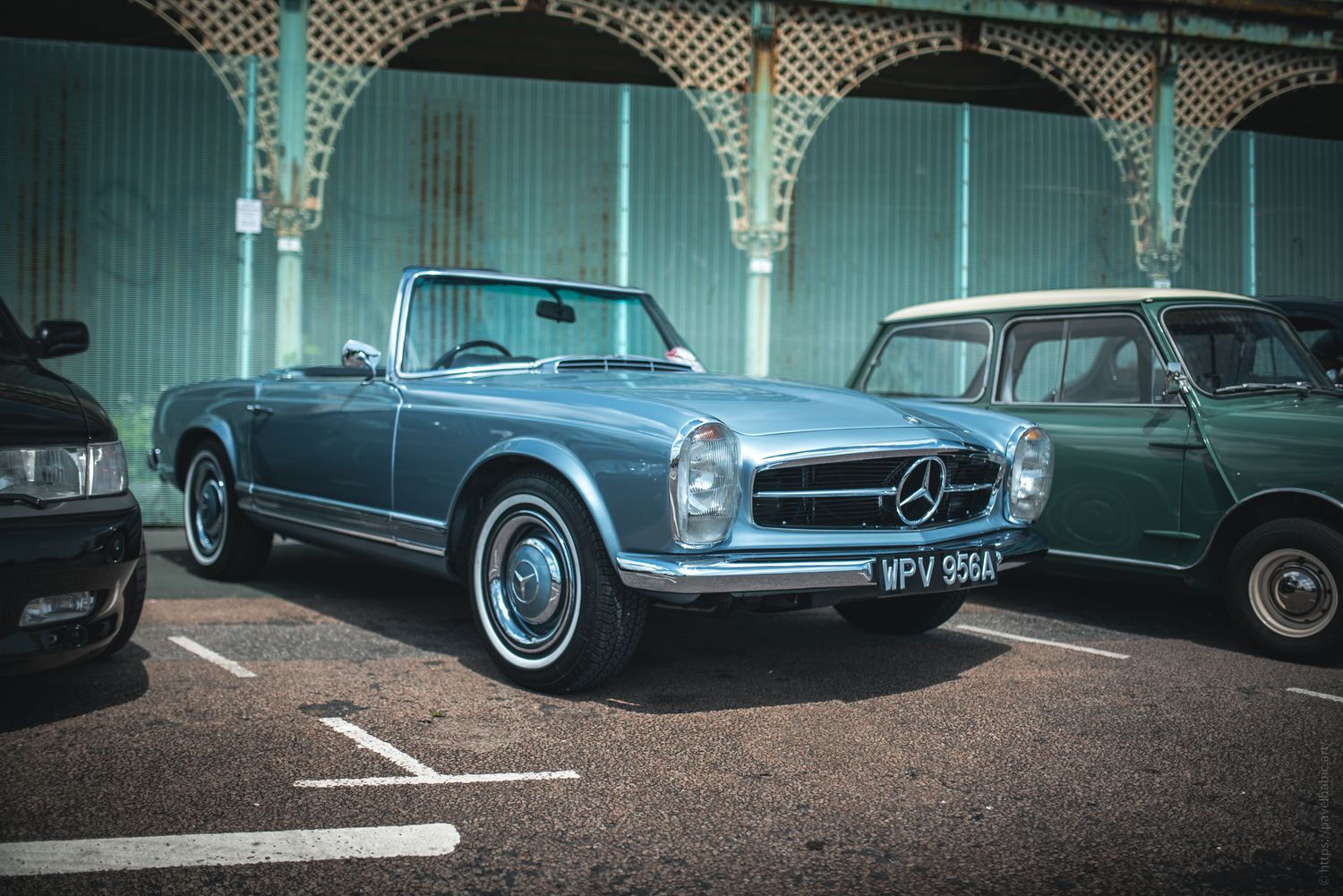 A classic blue Mercedes-Benz convertible parked between two vintage cars, set against a backdrop of an arched green wall.
