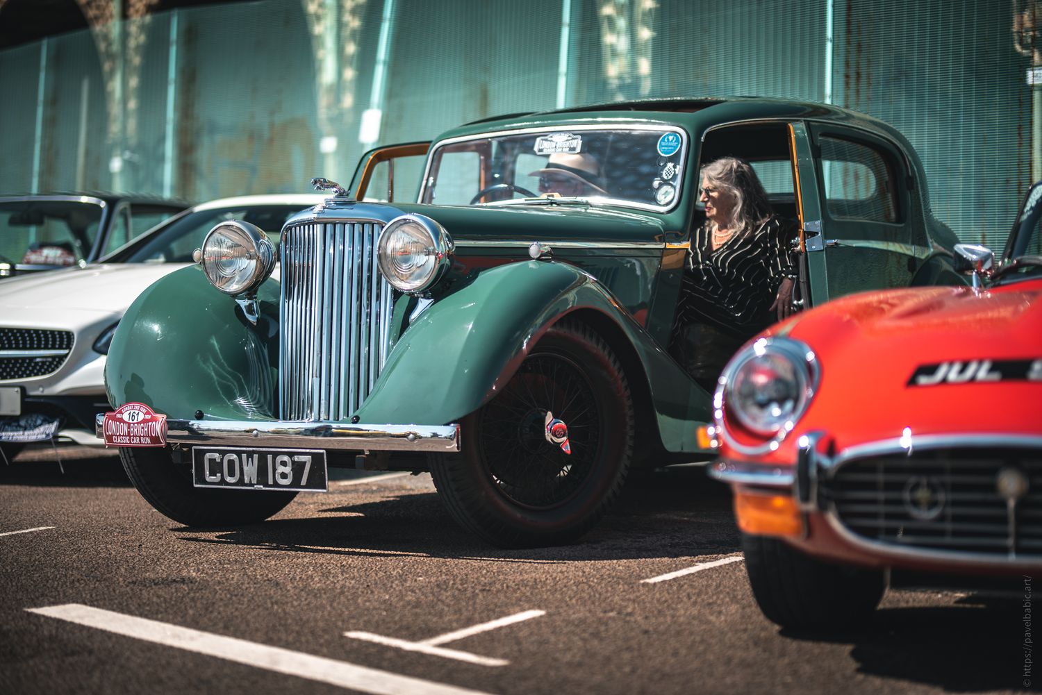 A vintage green car parked alongside modern vehicles, with a woman sitting inside, enjoying the sunny day.