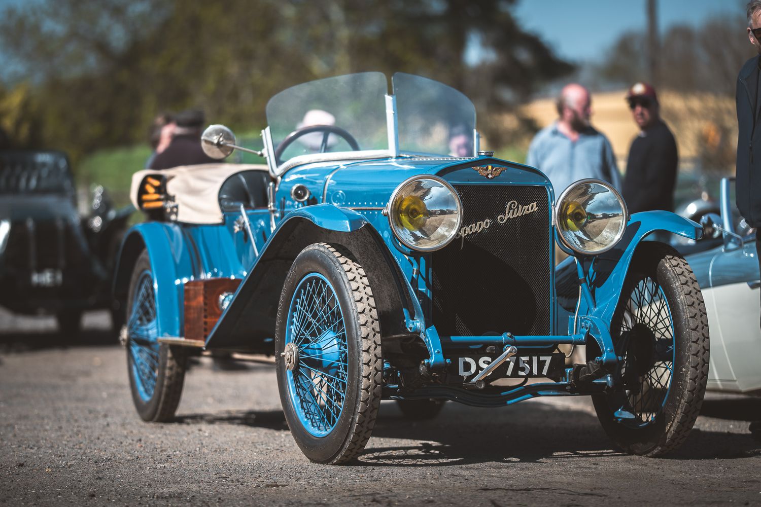 A vintage blue automobile with a stylish design, featuring wire wheels and a clear windshield, surrounded by people at a car event.