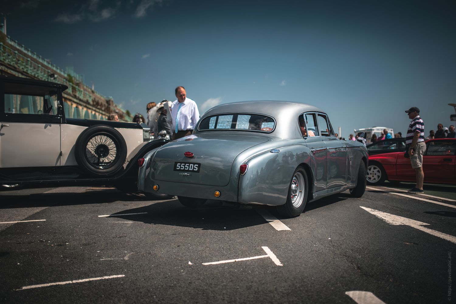 A sleek silver vintage car parked amid classic cars and spectators on a sunny day.