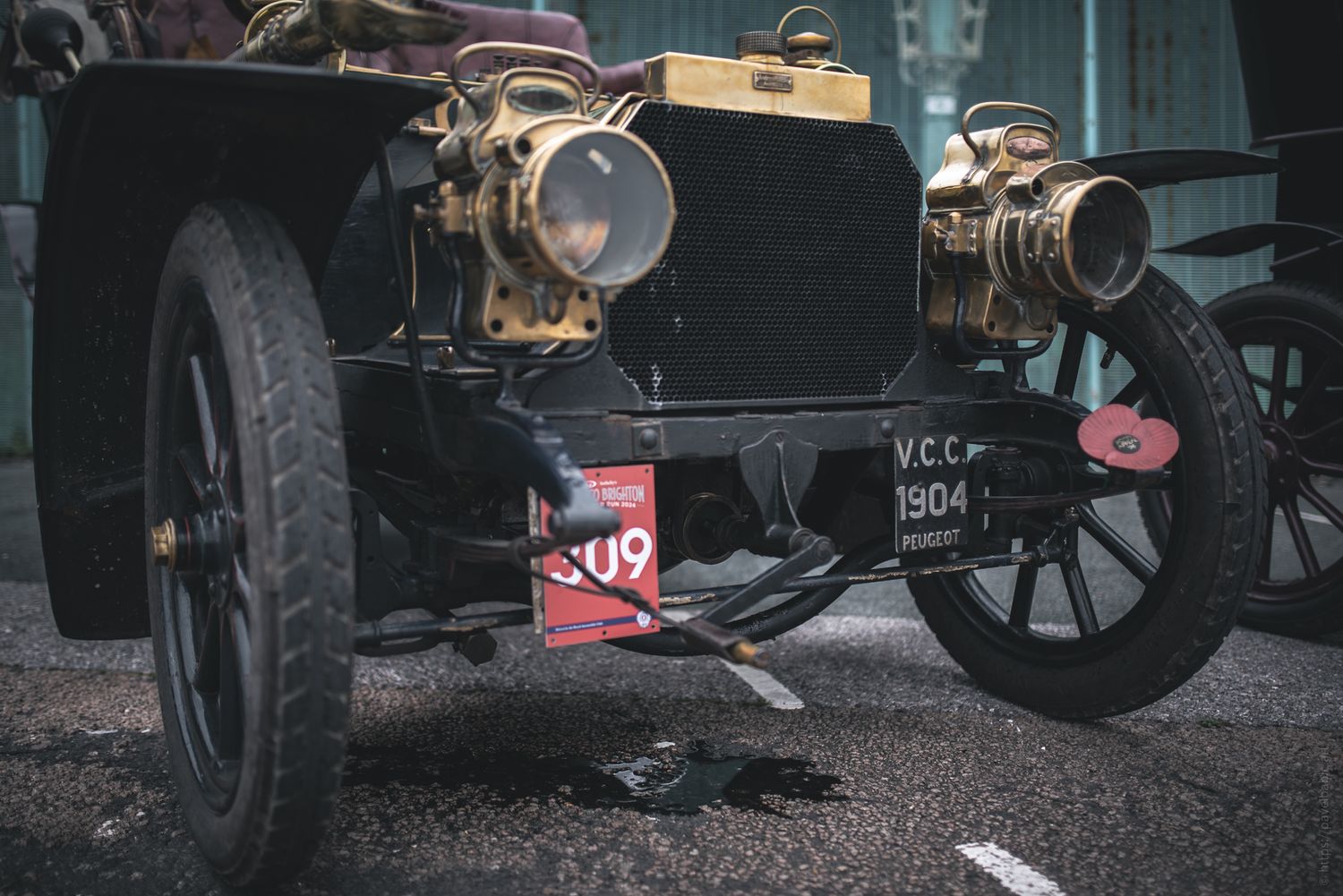 Close-up of a vintage 1904 car with brass headlights and a red license plate, showcased outdoors. The vehicle has a classic black chassis.