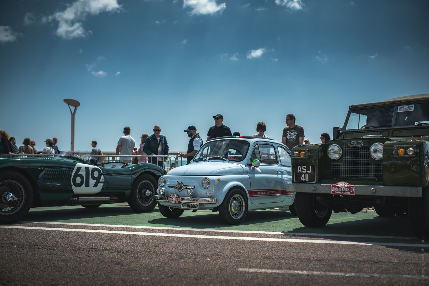 A vintage car show featuring a light blue Fiat, a dark green Land Rover, and classic race cars, with spectators enjoying the event.
