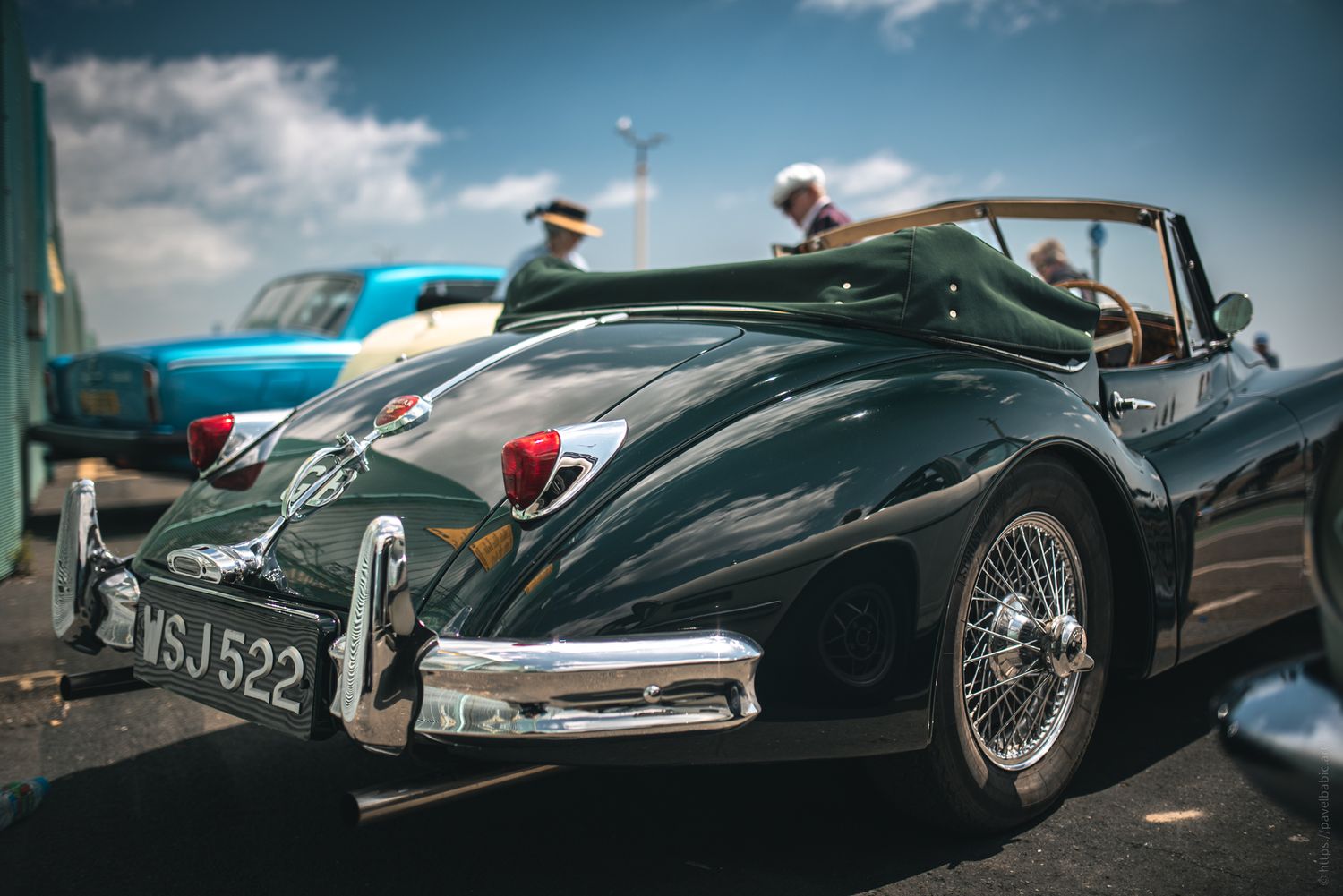 A classic black convertible car with chrome details parked, featuring a tan leather interior and a scenic backdrop with clouds.