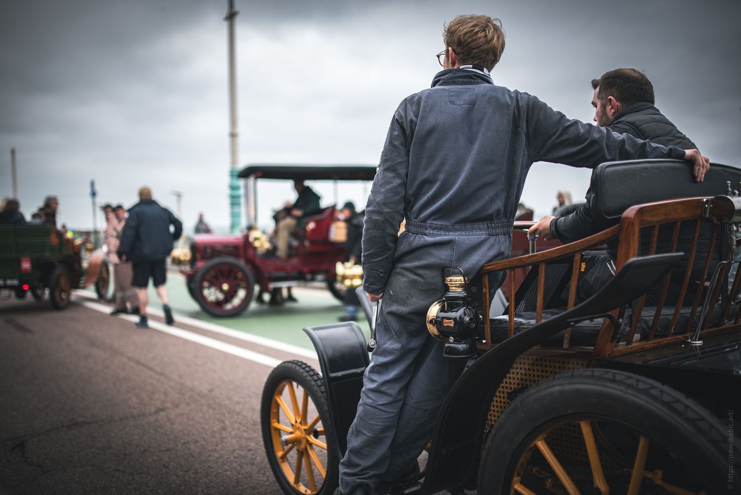 Two men in vintage attire admire classic cars at a gathering, with cloudy skies overhead and historical vehicles in the background.