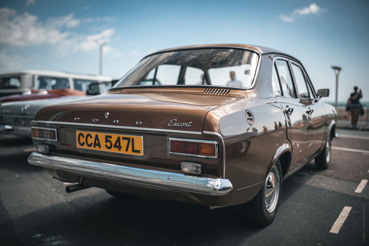 A classic brown Ford Escort parked, showcasing its vintage design, rear view, and license plate CCA 547L against a blue sky.