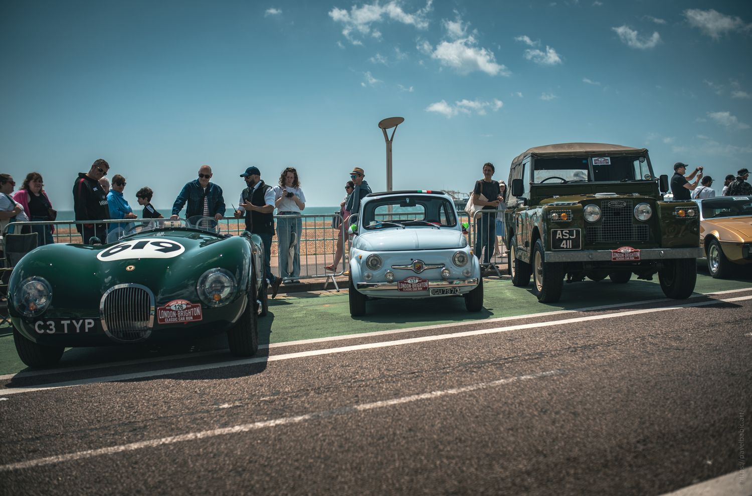 Three classic cars parked along a sunny boulevard, with a crowd of onlookers admiring them under a clear blue sky.