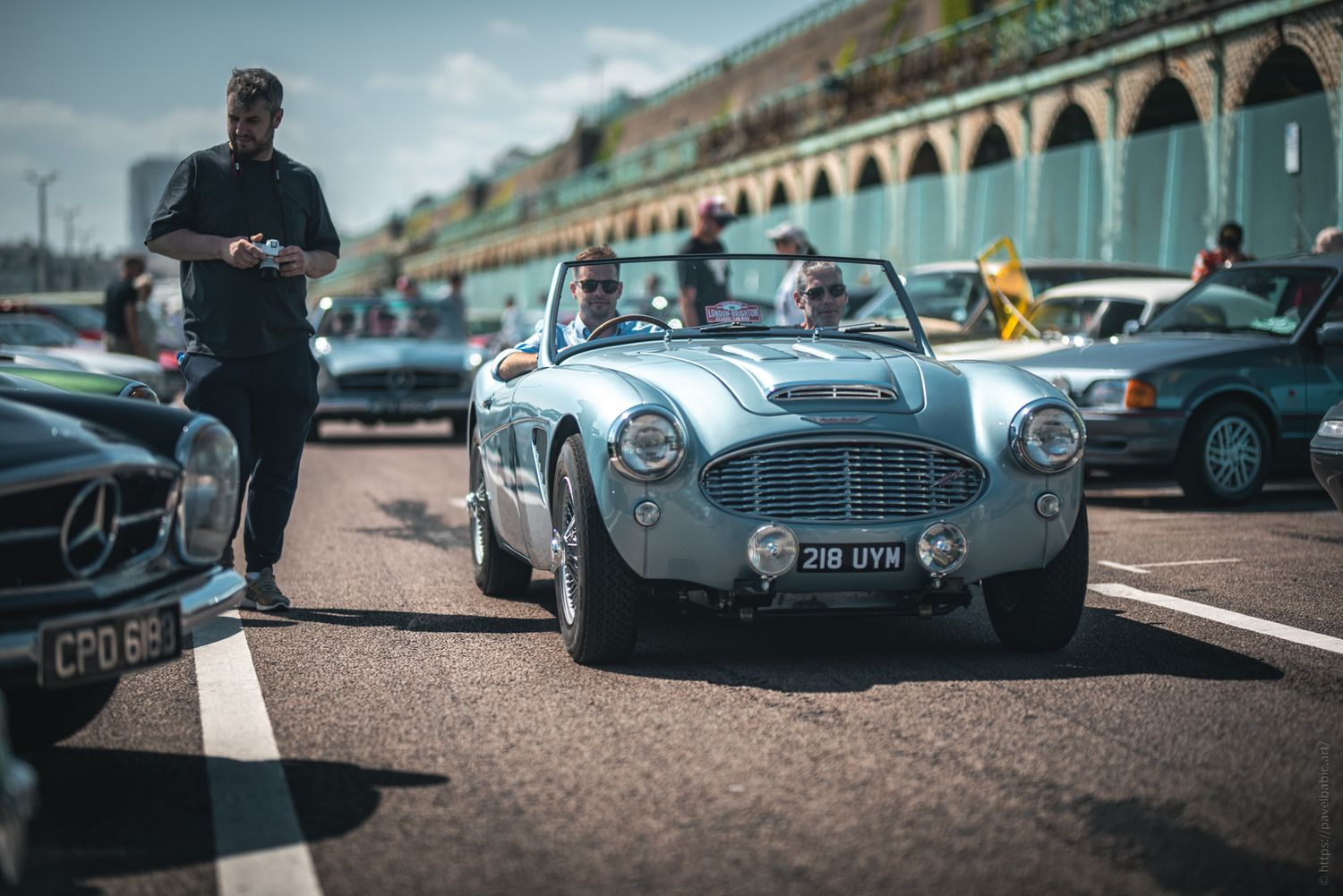 A classic convertible car drives along a seaside road with spectators nearby, showcasing vintage vehicles at a car show.