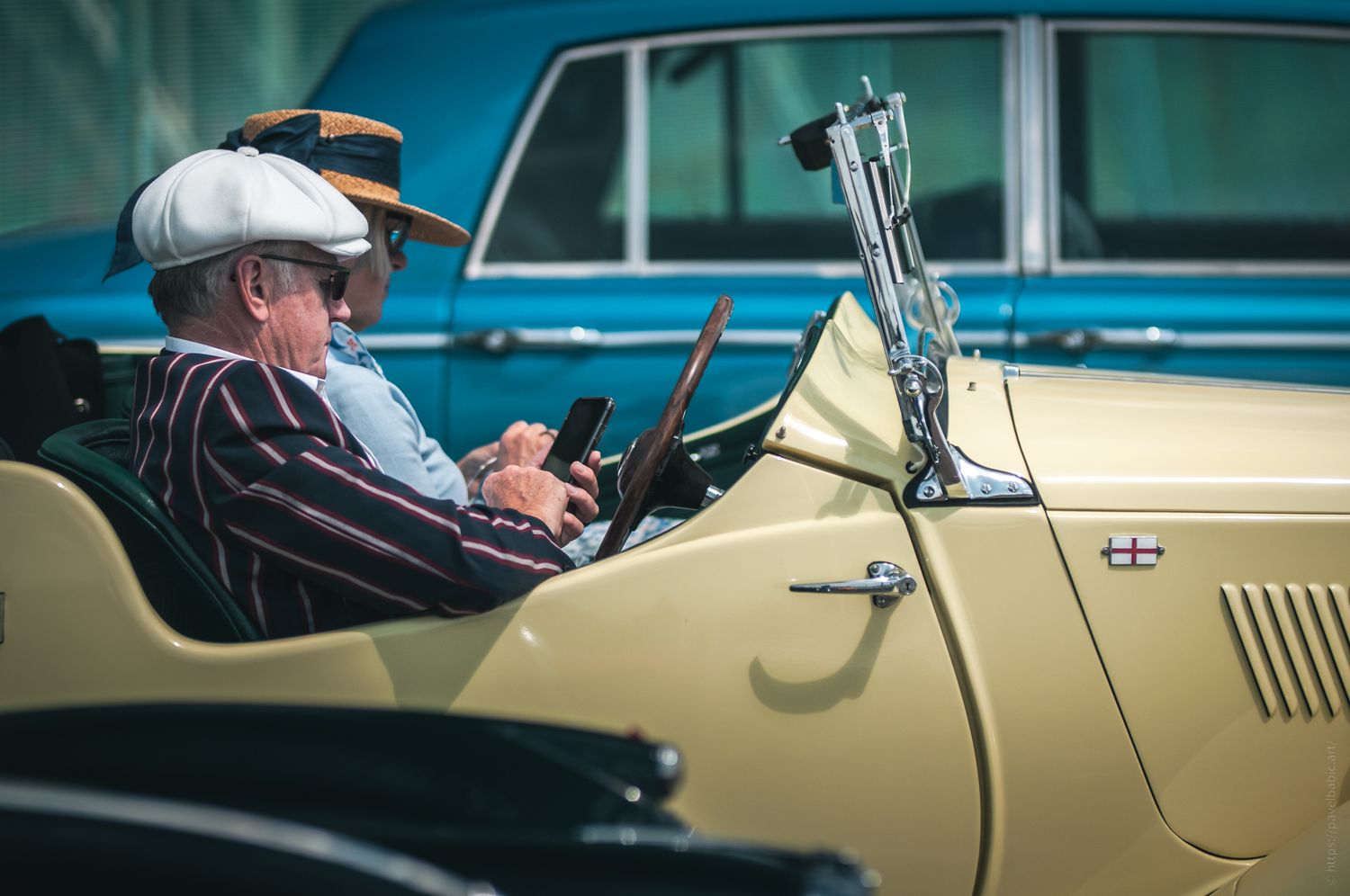 Two men sit in a vintage yellow convertible car, one using a smartphone while wearing a white cap and the other in a straw hat.