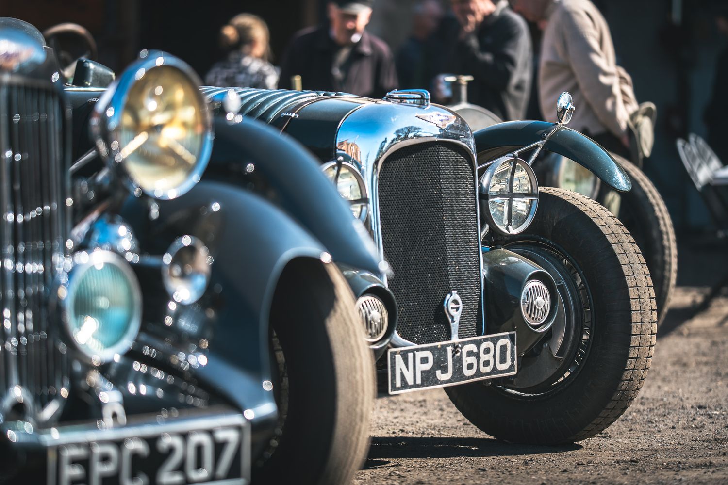 Vintage cars lined up at an event, showcasing their polished chrome details and classic designs, with spectators in the background.