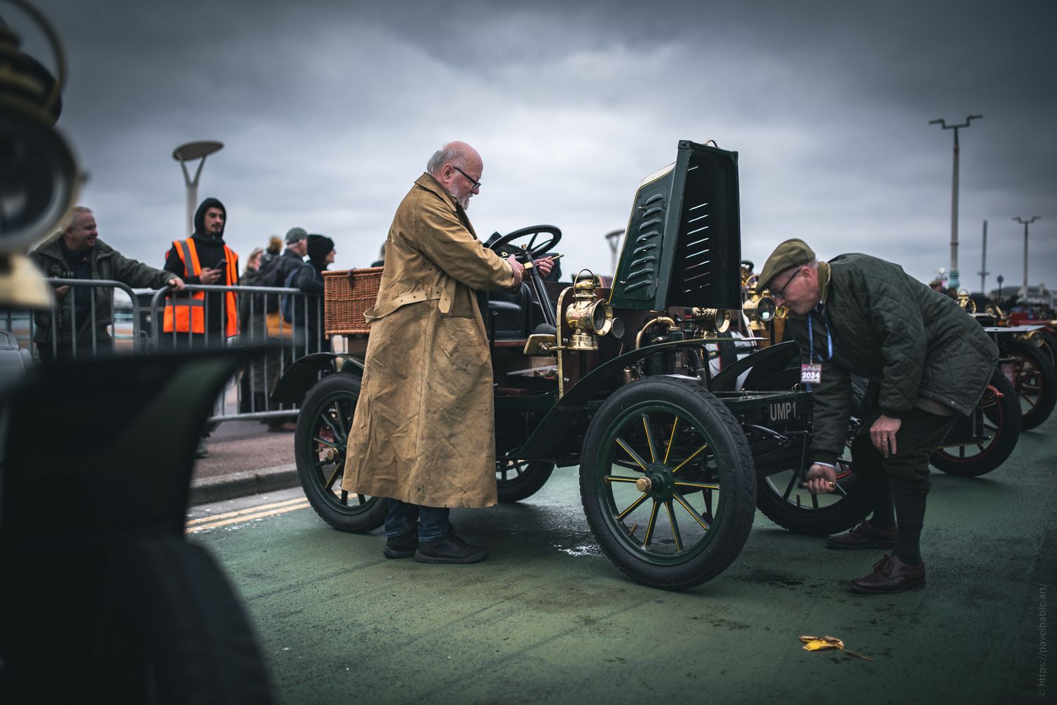 Two men in period attire work on a vintage car at an outdoor event, with onlookers in the background under a cloudy sky.