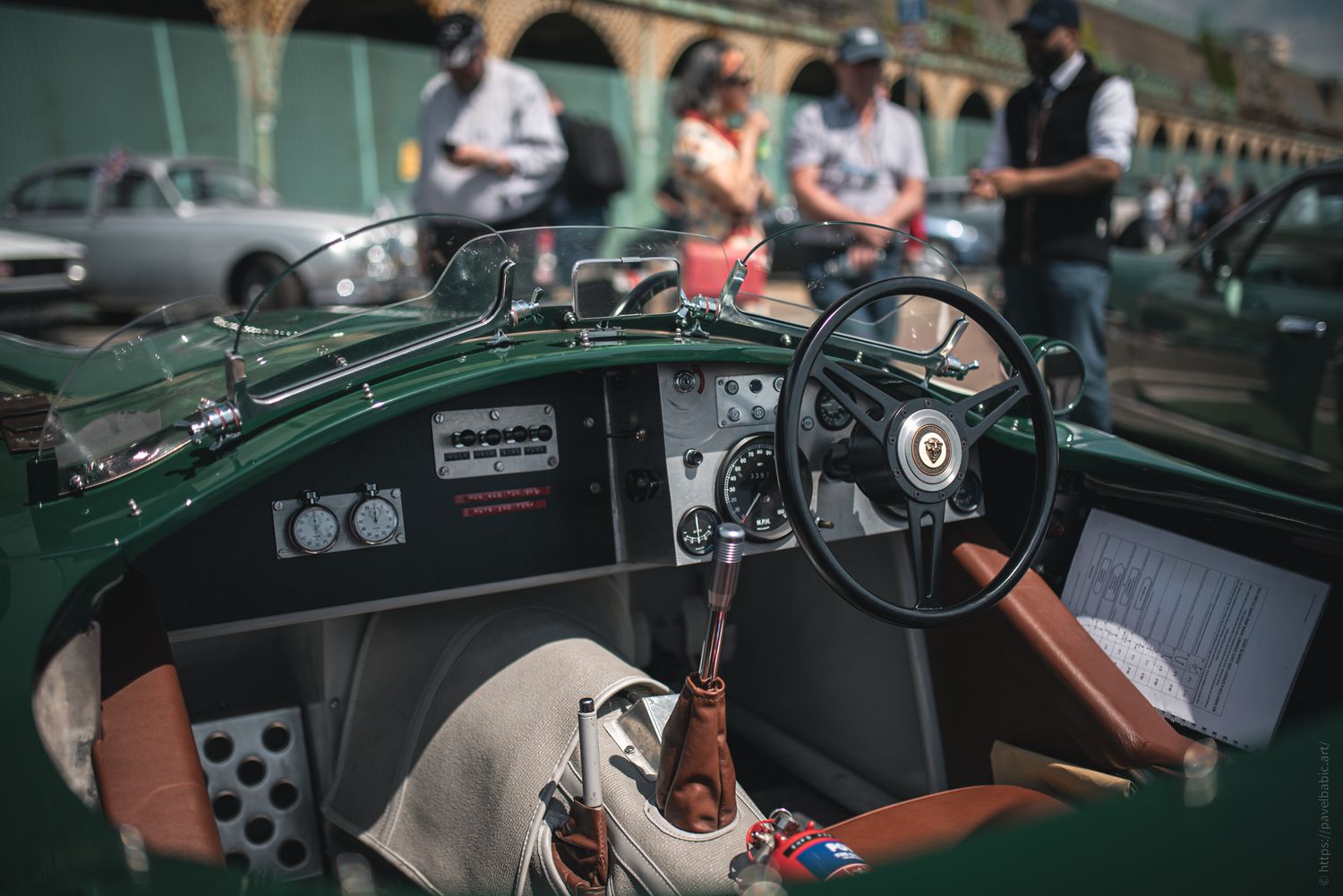 Close-up of a vintage green car's interior, featuring a leather seat, steering wheel, and gauges, with people admiring cars in the backgroun