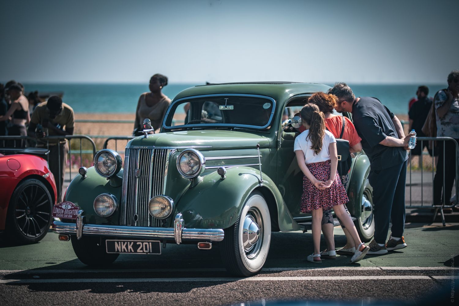 A vintage green car at a seaside event, with people admiring it, including a girl in a red skirt. A lively atmosphere surrounds them.