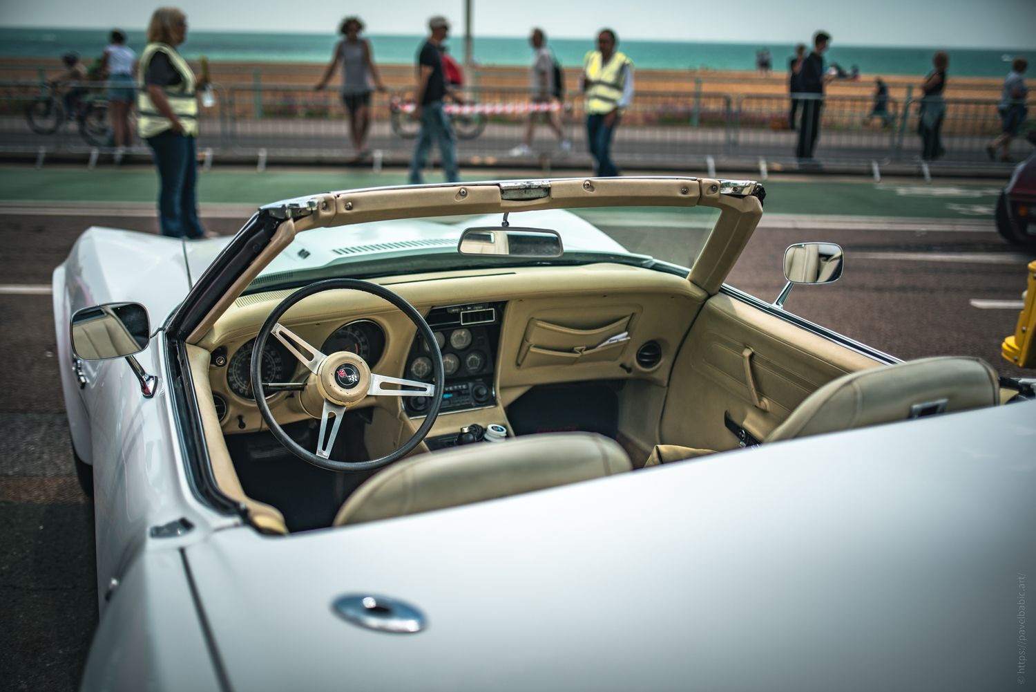 View of a classic convertible car's interior, with a beach setting and people walking in the background.