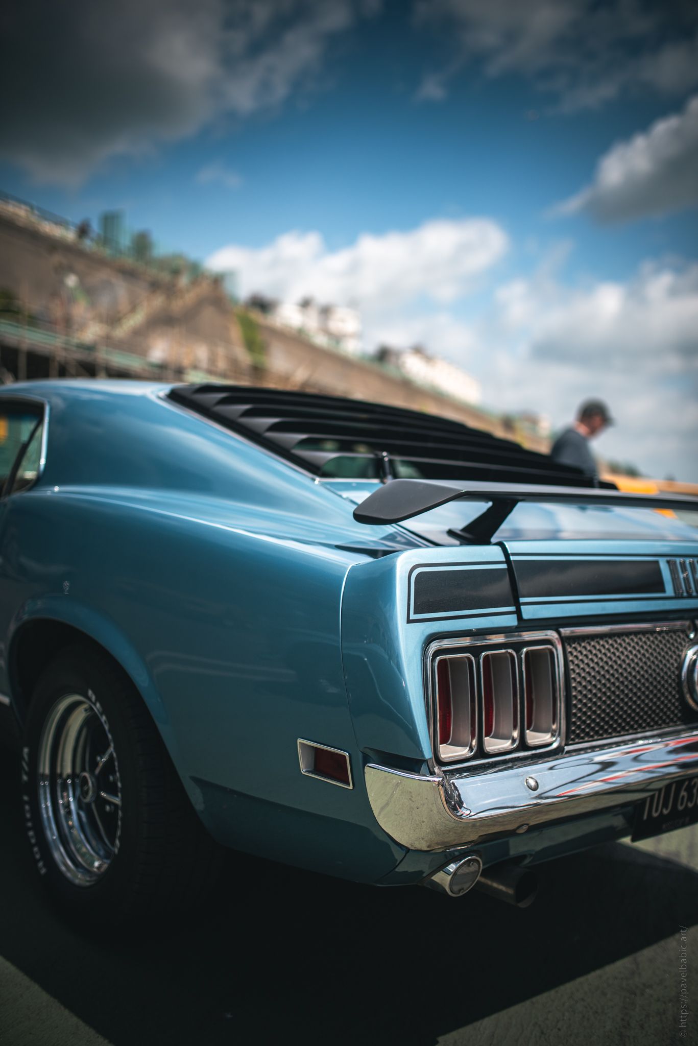 A classic blue muscle car with a sleek rear design, chrome details, and a prominent spoiler, set against a cloudy sky.