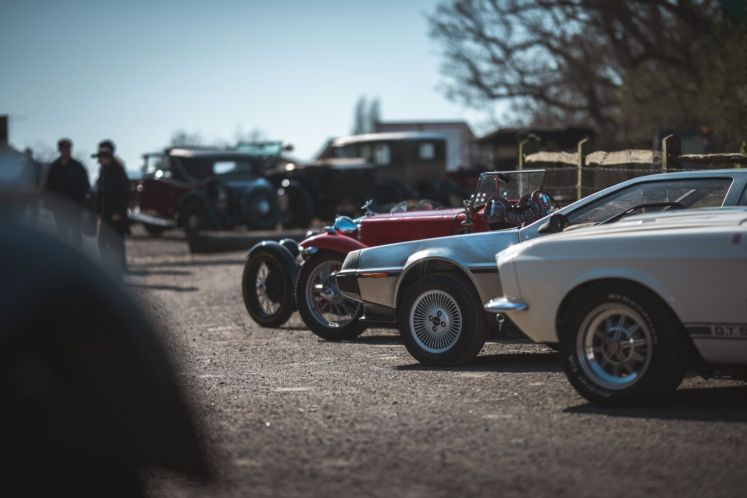 A row of vintage and classic cars parked on a gravel path, with spectators in the background enjoying a car show.