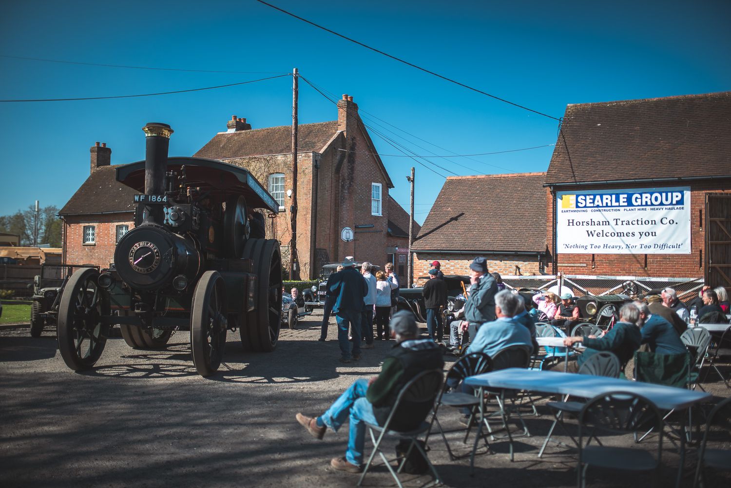 Steam traction engine at a rustic event with seated onlookers outside a brick building under clear blue skies; conveys a nostalgic