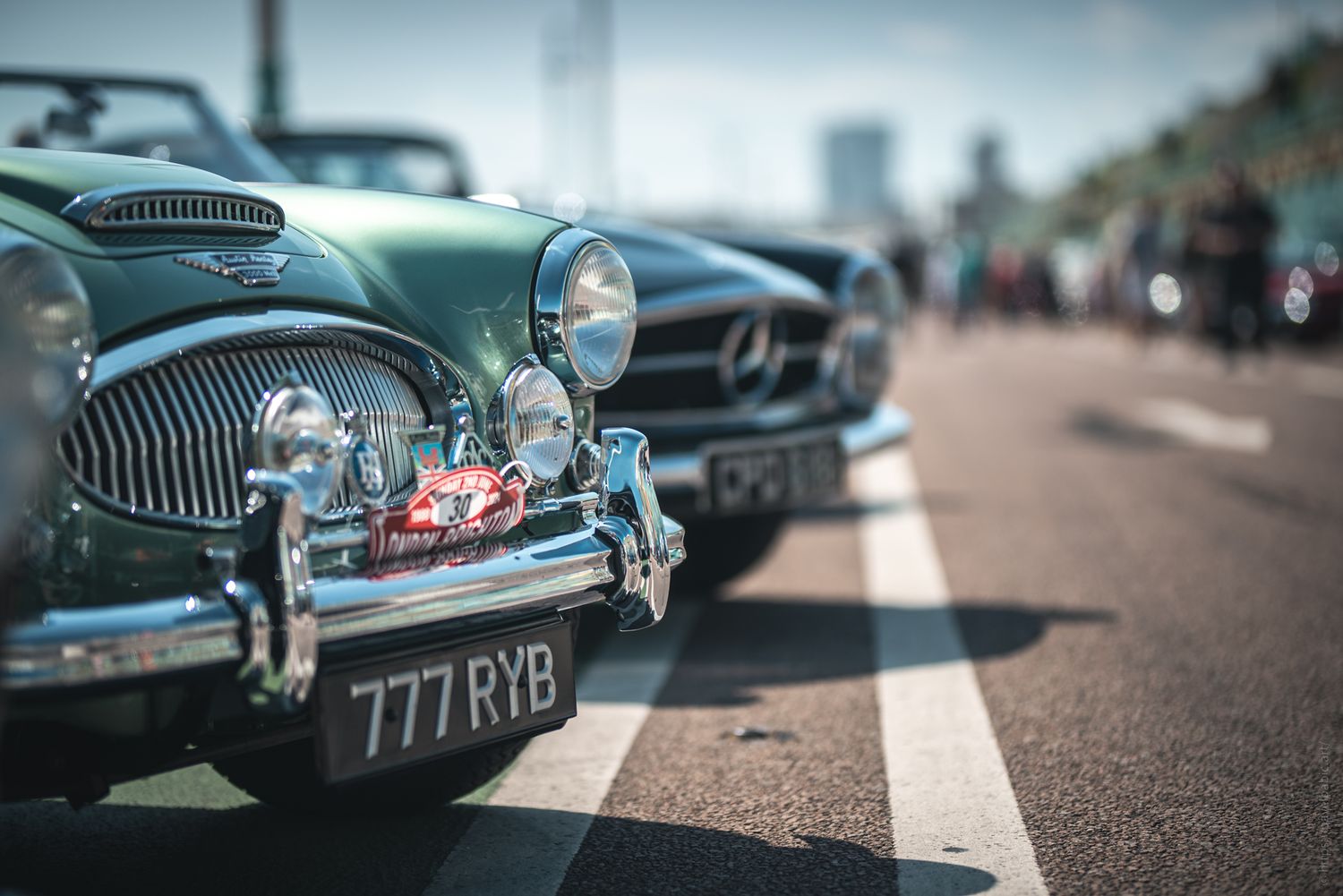 A close-up of vintage cars, including a green classic and a sleek Mercedes, lined up on a sunny street.