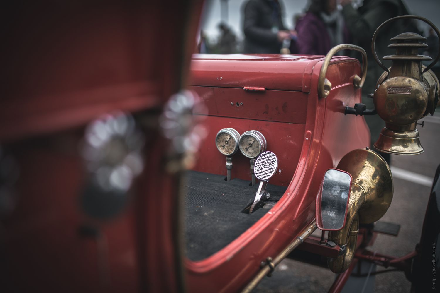 Close-up of a vintage red car’s dashboard featuring classic gauges and a brass lamp, set against a crowd in the background.