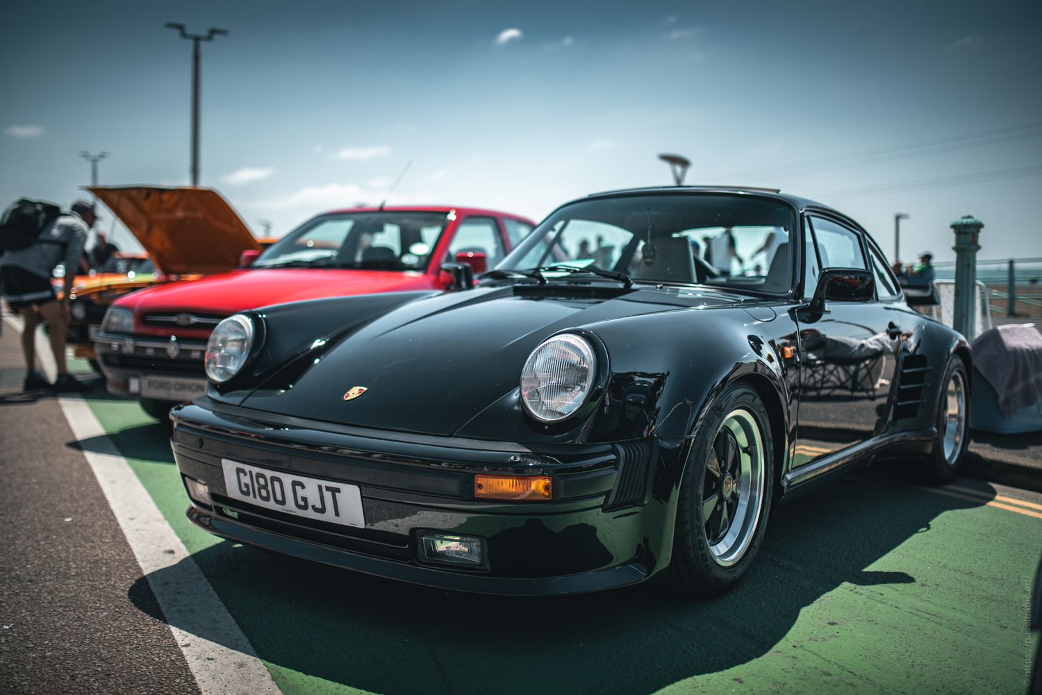 A sleek black Porsche sports car parked alongside a red car, set against a sunny sky and urban backdrop.