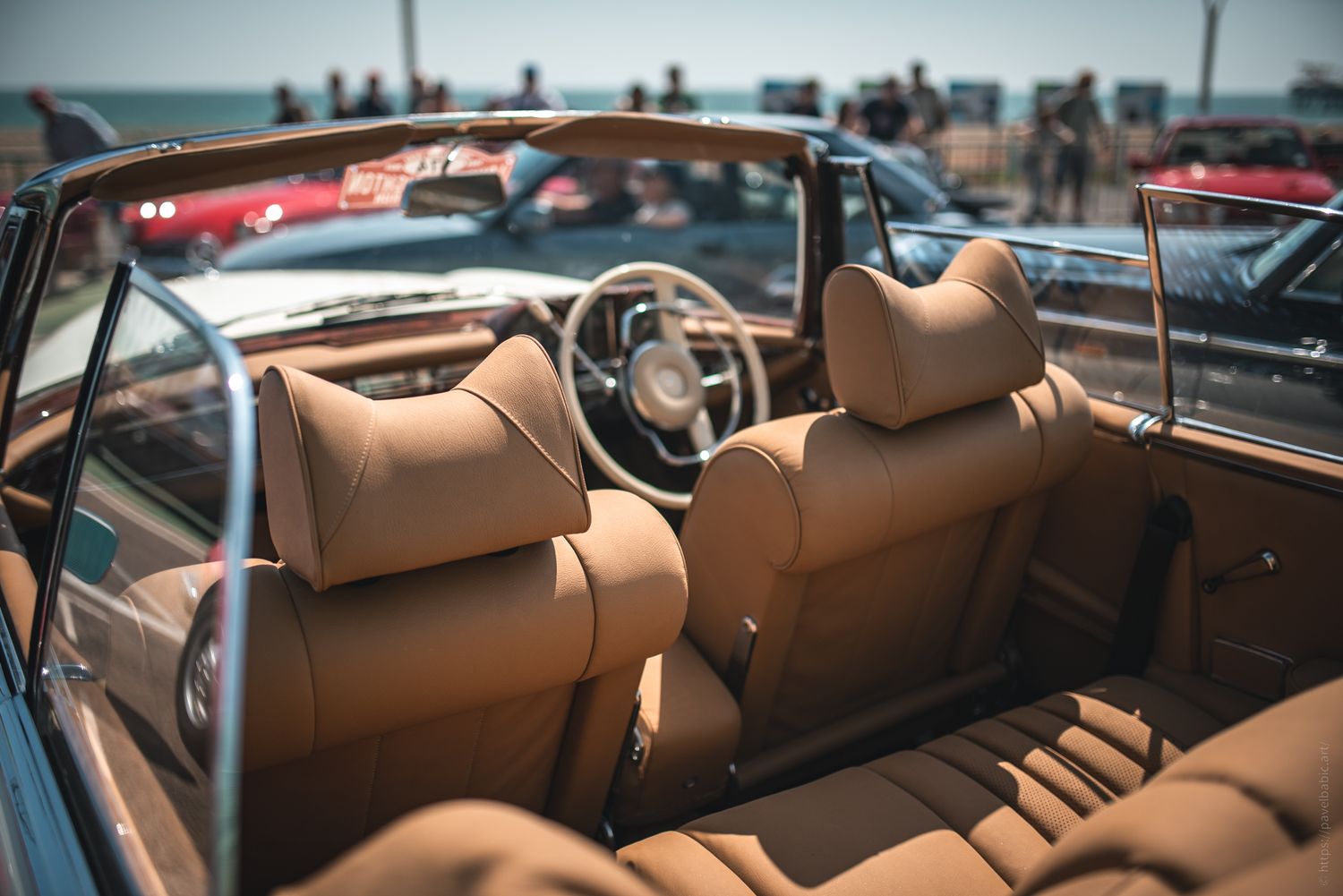 Interior of a stylish convertible car featuring tan leather seats, a classic steering wheel, and a vibrant outdoor crowd in the background.