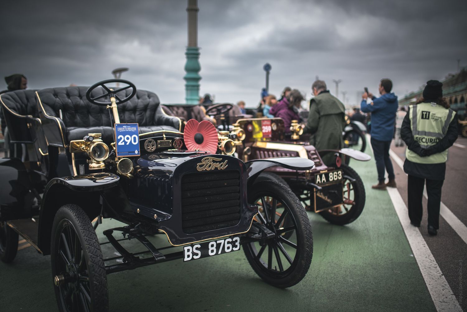 Two vintage cars with brass details and poppies lined up at a seaside event, with spectators and cloudy skies in the background.